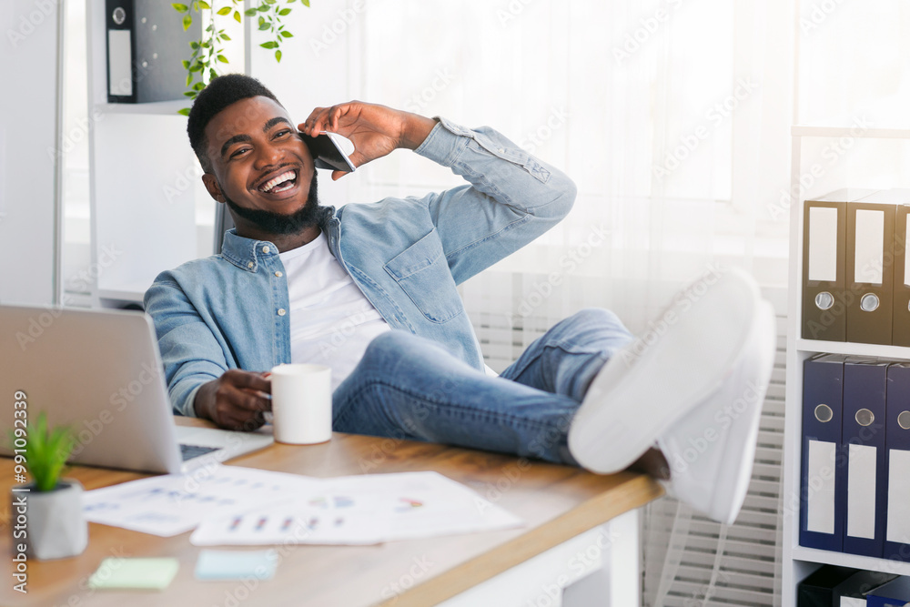 Poster joyful black manager talking on phone and drinking coffee while having break at work, leaning back o