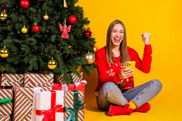 Full size photo of pretty young woman use smart phone raise fist tree wear christmas sweater isolated on yellow color background