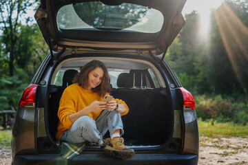 Young cheerful woman traveling by car taking a selfie on a smartphone while sitting in car open...
