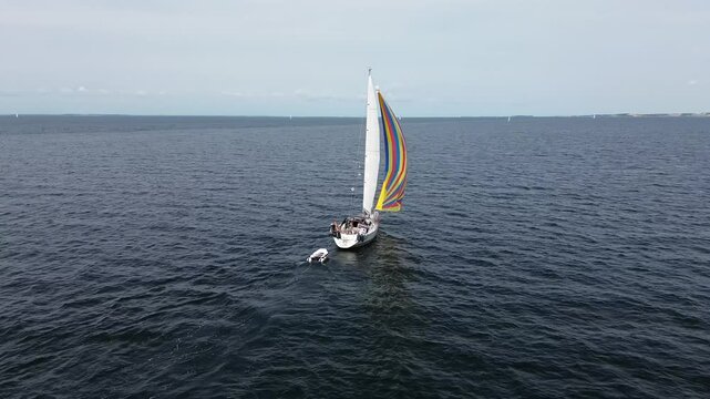 Aerial drone 4K film of sailboat yacht with colorful spinaker sail sailing on sea ocean water with Dutch holland flag on Baltic sea in front of lighthouse
