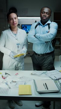 Vertical portrait shot of diverse detective and forensic scientist posing together for camera during criminal investigation in laboratory