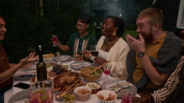 Slowmo of five multiracial friends laughing and raising glasses in toast while sitting at festive dinner table in backyard on summer evening
