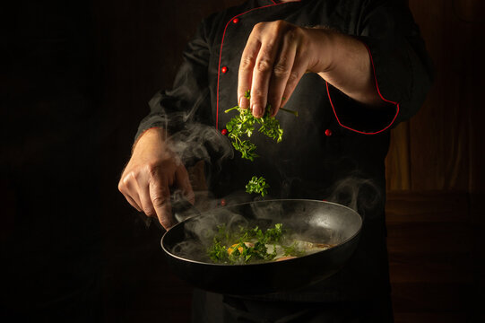 The chef adds aromatic parsley with his hand to a steaming hot frying pan with fried eggs. Low key concept of preparing breakfast in the kitchen with black advertising space. Thai cuisine