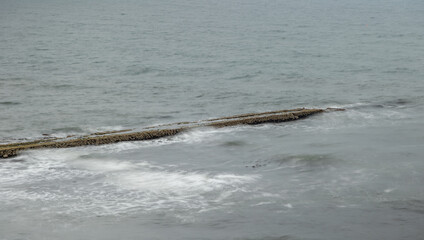 Fototapeta premium Long exposure of granite stone pier seawall and rocks along coast coastline in England, uk, Newcastle with blurred waves and grey water of the sea ocean