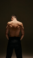 Fototapeta premium Rear view of young man posing shirtless in pants, showing his perfect, sportive body, relief strong back against dark studio background. Concept of male beauty, body and skin care, health, sport