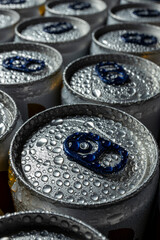 Close-up of multiple aluminum beverage cans arranged in rows, covered in condensation. The blue pull tabs and water droplets on the metallic surface emphasize coldness and freshness.