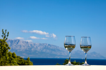 Two glasses of wine filled with white wine stand against the backdrop of mountains, white clouds, the Adriatic Sea and a clear, bright, blue sky in Croatia in the summer.