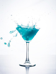Blue cocktail in martini glass with ice cube splashing into liquid against white background. Blue curacao cocktail with splash on white.
