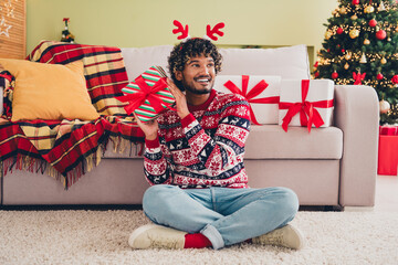 Full size photo of nice young man sit floor shake giftbox wear christmas pullover festive time apartment indoors