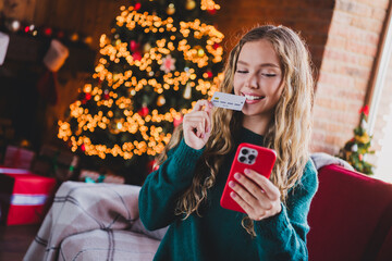 Photo of cheerful teen girl eshop device bite credit card dressed green christmas garment tradition atmosphere decor house indoors room