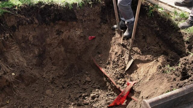 Workers use shovels to dig a deep hole in a backyard surrounded by grass and soil. The sun casts shadows as they aim for precision in their excavation efforts, showing dedication to completing the tas