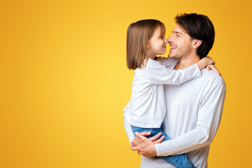 Portrait of sweet little girl looking at her smiling dad, father holding daughter over yellow background, copy space
