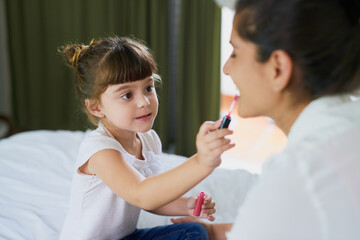 Mom, girl child and lipstick in bedroom with brush, smile or helping hand with beauty in family home. Cosmetics, mother and daughter with color, mouth and makeup for learning with bonding in house