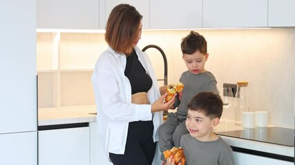 mom and sons sibling in kitchen in the morning eating croissant with chocolate,drinking tea. baby boy sitting on counter,mommy washing glass,sink in background.woman hugging kids, happy family