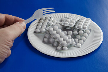 hand holding a fork over a white plate with white tablets in blisters on a general blue background, close-up, daily dose of medication, taking medication, large dose of drugs