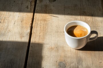 Obraz premium A white cup of espresso sitting on a rustic wooden table, with natural light casting a soft shadow over the surface, evoking a warm and cozy atmosphere.