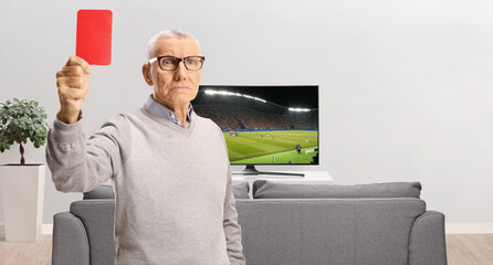 Serious elderly man showing a red card in front of tv screen showing a football match