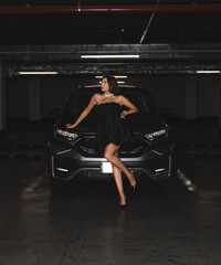 attractive woman posing in front of modern suv car in underground parking.female in dress painting lips in rear view mirror of auto vehicle.smiling lady in black dress with legs outside from window.