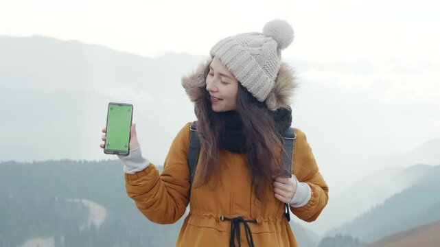 Happy asian woman tourist portrait show green screen mobile phone high in cloudy mountains at cold weather. Young hiker at background of mountain landscape in smoke. Copy space chroma key application.