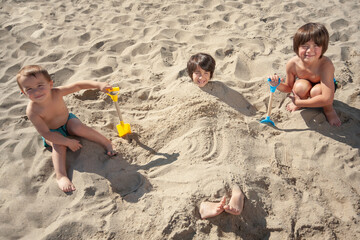Little brothers pose contentedly after burying their older brother in the sand on the beach with...