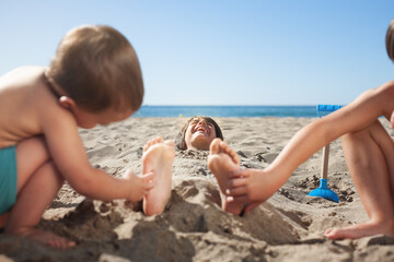 Child buried in the sand on the beach laughs when his siblings tickle his feet.