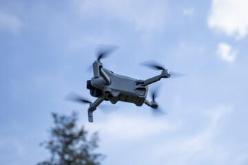 A flying drone captured mid-air against a clear blue sky with motion blur on the propellers. The background includes a blurred tree, suggesting the drone is being used outdoors.