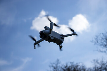 A flying drone captured in mid-air against a blue sky with soft clouds. The drone is in focus, showcasing its propellers in motion, with a blurred backdrop of trees.