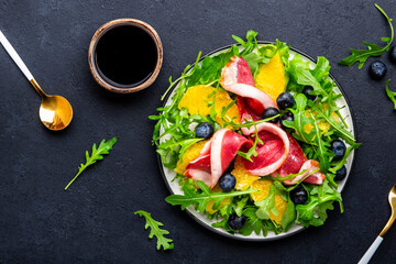 Foodie food salad with raw smoked duck and juicy orange, blueberry and arugula on black background, top view