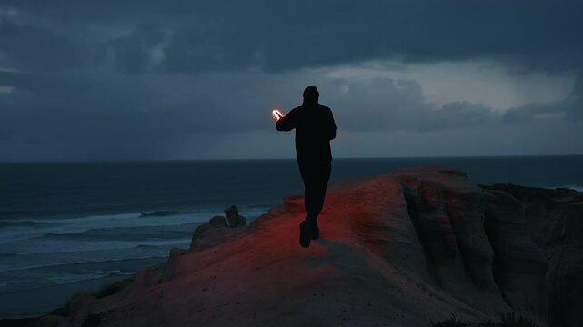 Man running up the coastline hill with red lit torch at dusk after sunset. Man with hat and face bandada at night with red burning signal flare in his hand. Young man with lit torch by the ocean.