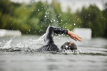 Side view of strong man swimming in forest lake with water splashes and wearing wet suit practicing...