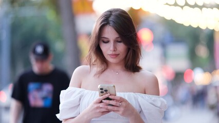 Carefree beautiful woman holding mobile phone, looking at mobile phone screen, using smartphone apps, posing outdoors.