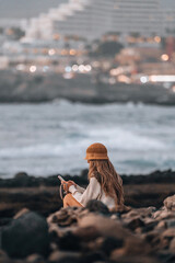 A woman is sitting on a rock by the ocean, looking at her phone