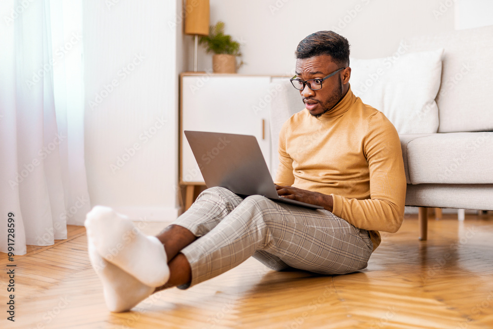 Poster shocked african american man using computer reading news on laptop sitting on floor at home. selecti