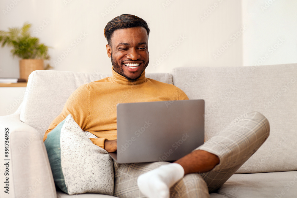 Poster Cheerful Black Man Using Laptop Playing Computer Games Sitting On Couch At Home. Selective Focus, Copy Space