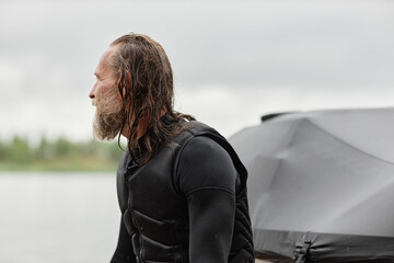 Minimal side view portrait of man wearing wetsuit and looking away sitting on back of boat in nature with long wet hair copy space