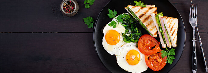 Breakfast: fried egg, spinach, tomatoes and club sandwich on plate. Top view, banner