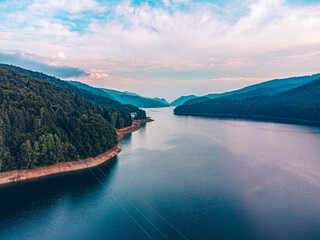 Drone perspective of a sunset over a mountain lake, with the water reflecting the colorful sky and surrounding hills.