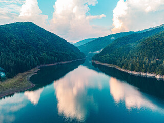 Drone view of a picturesque lake during sunset, bordered by rugged mountains and a colorful sky.
