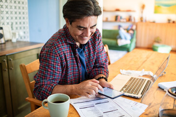 Smiling man using smartphone while working on laptop at home