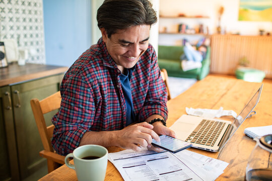 Smiling man using smartphone while working on laptop at home