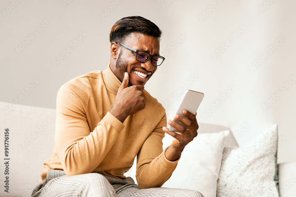 Fototapete joyful afro guy using cellphone texting or reading funny news on internet sitting on couch at home. 