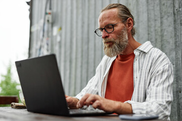 Fototapeta premium Portrait of bearded mature man wearing glasses and using laptop while working or using internet sitting at wooden table outdoors copy space