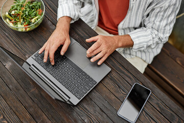 Top view closeup of unrecognizable adult man typing on laptop keyboard while working at table in outdoor cafe or terrace