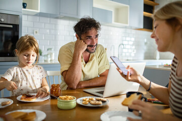 Father working on laptop while family has breakfast in kitchen