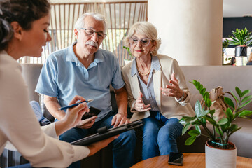 Obraz premium An elderly couple sits in a modern hotel lobby, talking with a hotel staff representative who is assisting them with the check-in process or presenting a hotel offer. Luggage is placed nearby.
