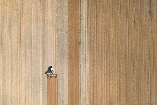 An aerial view of a tractor working on a large agricultural field, plowing the soil in straight, parallel rows.