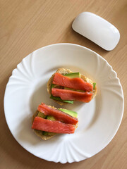 Fresh avocado and smoked salmon toast on white plate. Healthy breakfast standing on a wooden table with a computer mouse nearby.