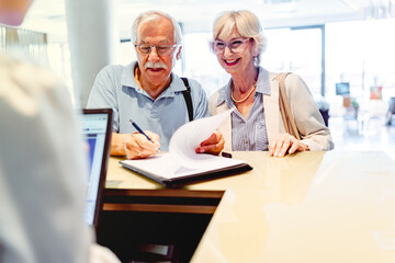 Obraz premium A smiling elderly couple stands in front of a hotel desk, filling out a check-in form or signing a bill, indicating they are either checking in or checking out after a wonderful stay.