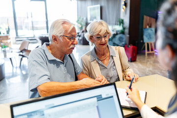 A smiling elderly couple stands in front of a hotel desk, filling out a check-in form or signing a...