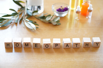 word HOMEOPATHY written on wooden blocks on a table with plants, herbs, natural oils and a stethoscope.